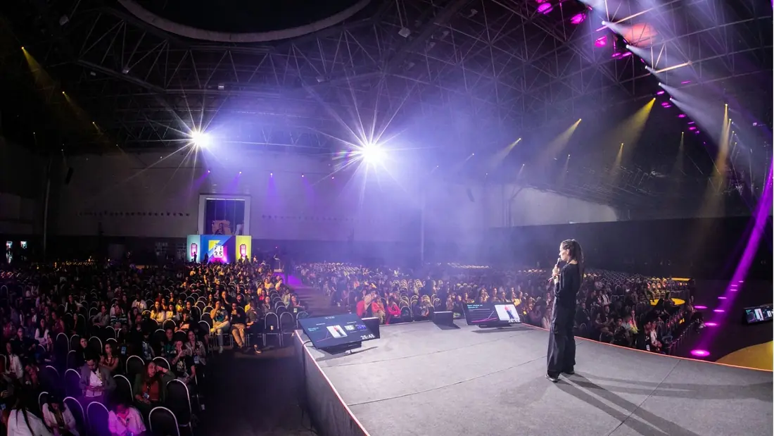 Monica Magalhaes at the microphone at the edge of the stage, facing a packed auditorium under purple light