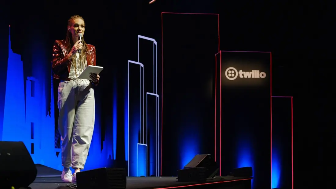 Monica Magalhaes on stage with a microphone and tablet, in front of a city-skyline silhouette backdrop